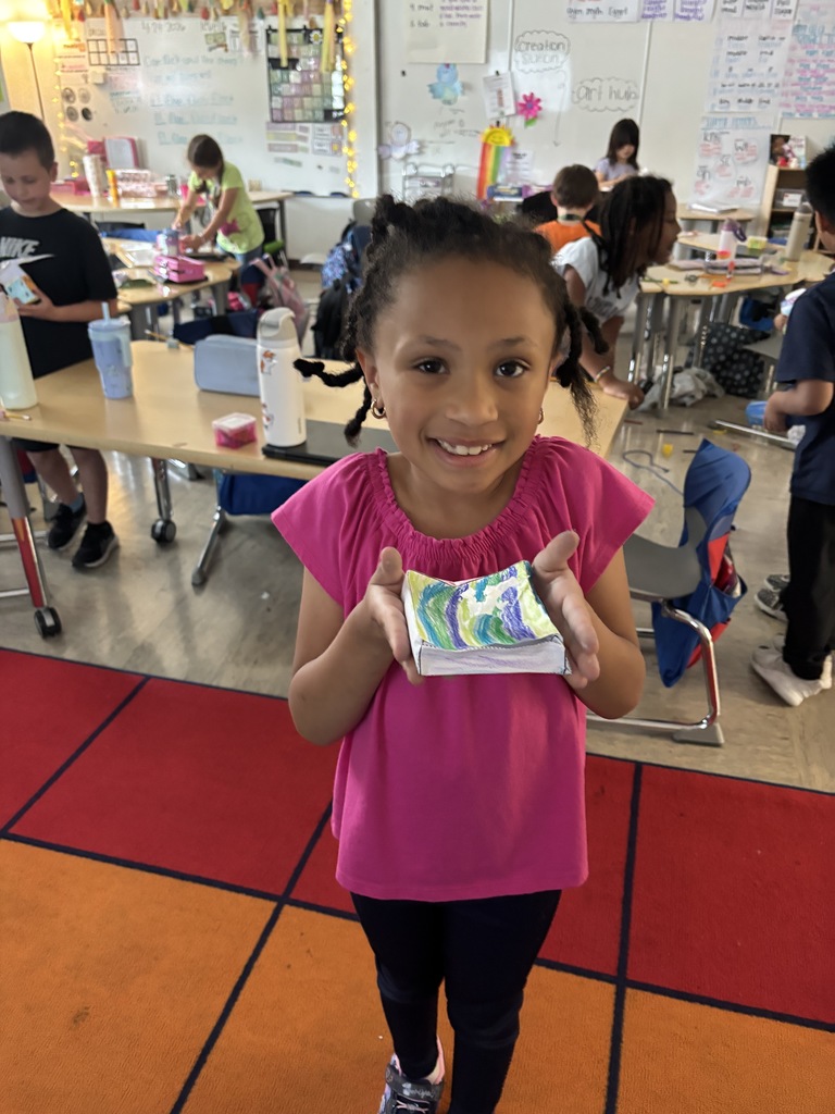 A student smiling and holding up a real-world example of a solid figure, demonstrating their understanding of 3D shapes during a hands-on math activity in the classroom.