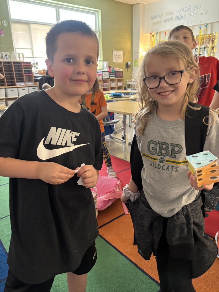 Two students smiling and holding up a real-world example of a solid figure, demonstrating their understanding of 3D shapes during a hands-on math activity in the classroom.