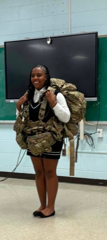 A smiling young woman standing in a classroom while wearing a large, heavy military rucksack and a tactical chest rig over her school uniform. The camouflage gear is bulky and oversized for her frame. She is standing in front of a green chalkboard and a large digital monitor.