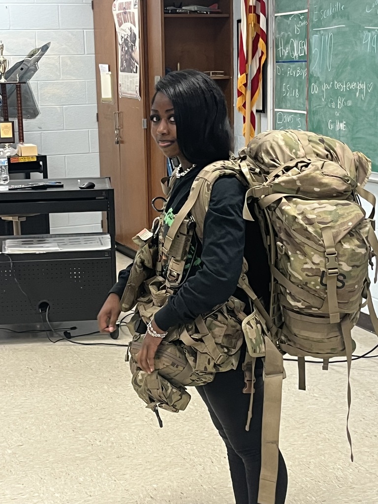 A young woman standing in a classroom wearing a massive, oversized military-style rucksack and tactical gear in OCP camouflage. The backpack is nearly as large as she is, extending from her head to her mid-thigh. She is looking toward the camera with a slight smile. In the background, there is a chalkboard with various notes and an American flag.