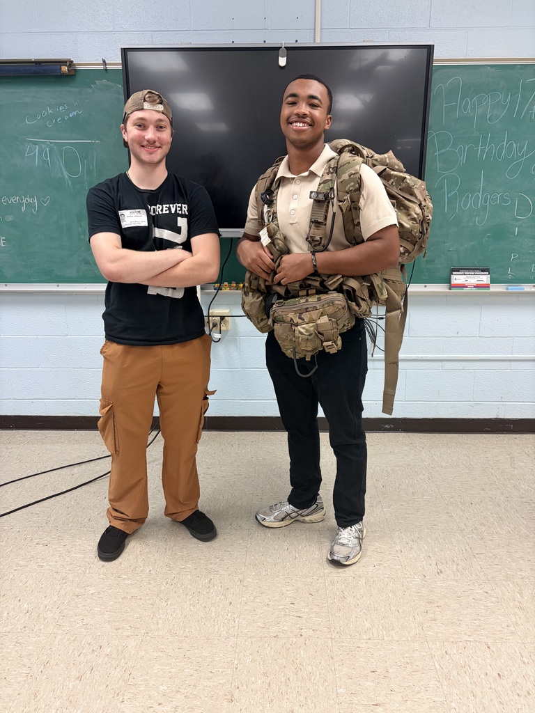 Two young men standing side-by-side in a classroom, smiling at the camera. The man on the right is wearing a large, camouflage military rucksack and tactical chest rig. The man on the left has his arms crossed and is wearing a baseball cap and a black t-shirt with a visitor badge. They are standing in front of a green chalkboard with handwritten notes and a large flat-screen monitor.