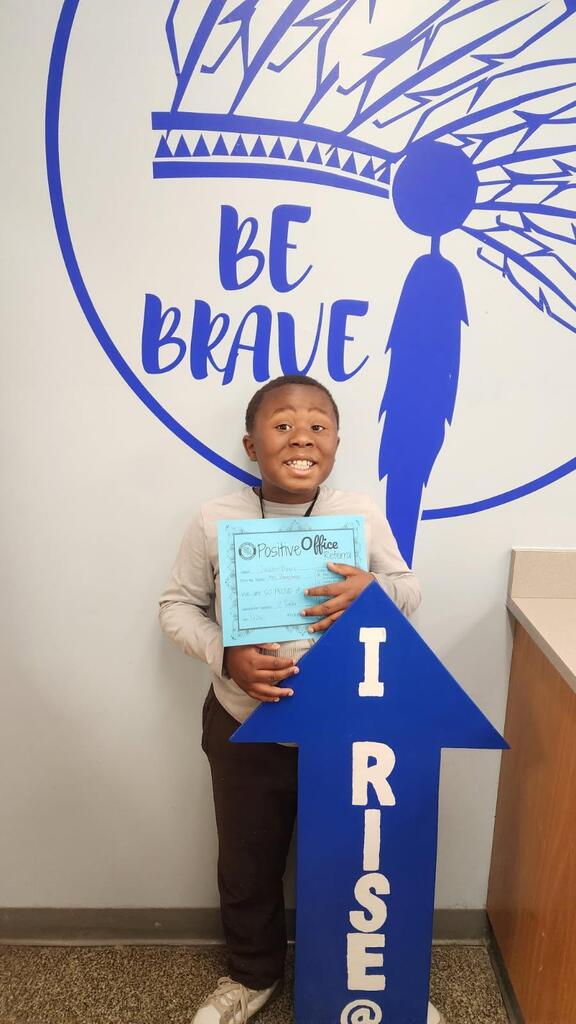 Student standing in the main office with the I RISE sign, proudly displaying their Positive Office Referral certificate.