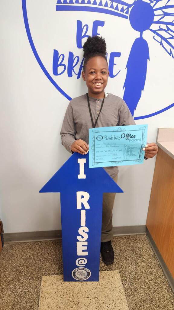Student standing in the main office with the I RISE sign, proudly displaying their Positive Office Referral certificate.
