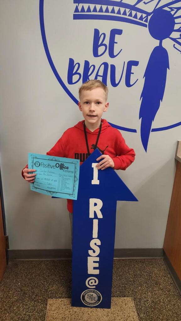 Student standing in the main office with the I RISE sign, proudly displaying their Positive Office Referral certificate.