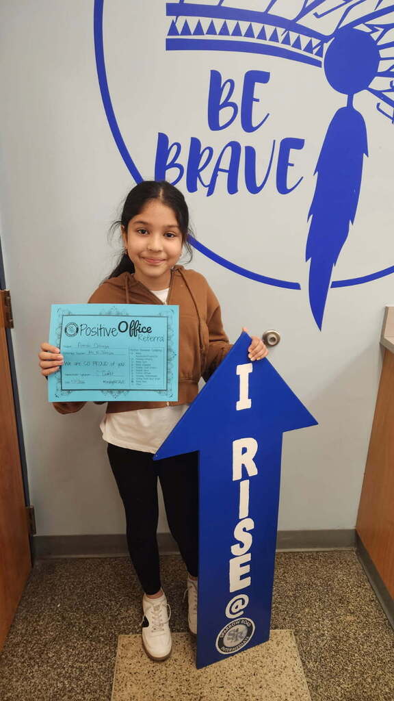 Student standing in the main office with the I RISE sign, proudly displaying their Positive Office Referral certificate.