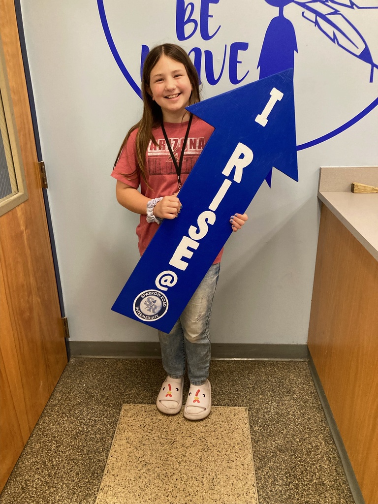 Student smiling and  standing in the main office proudly holding the I RISE sign.