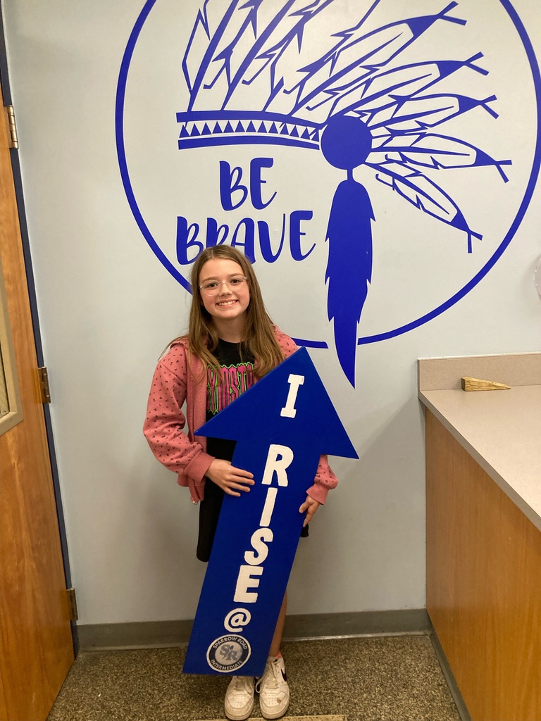 Student smiling and  standing in the main office proudly holding the I RISE sign.