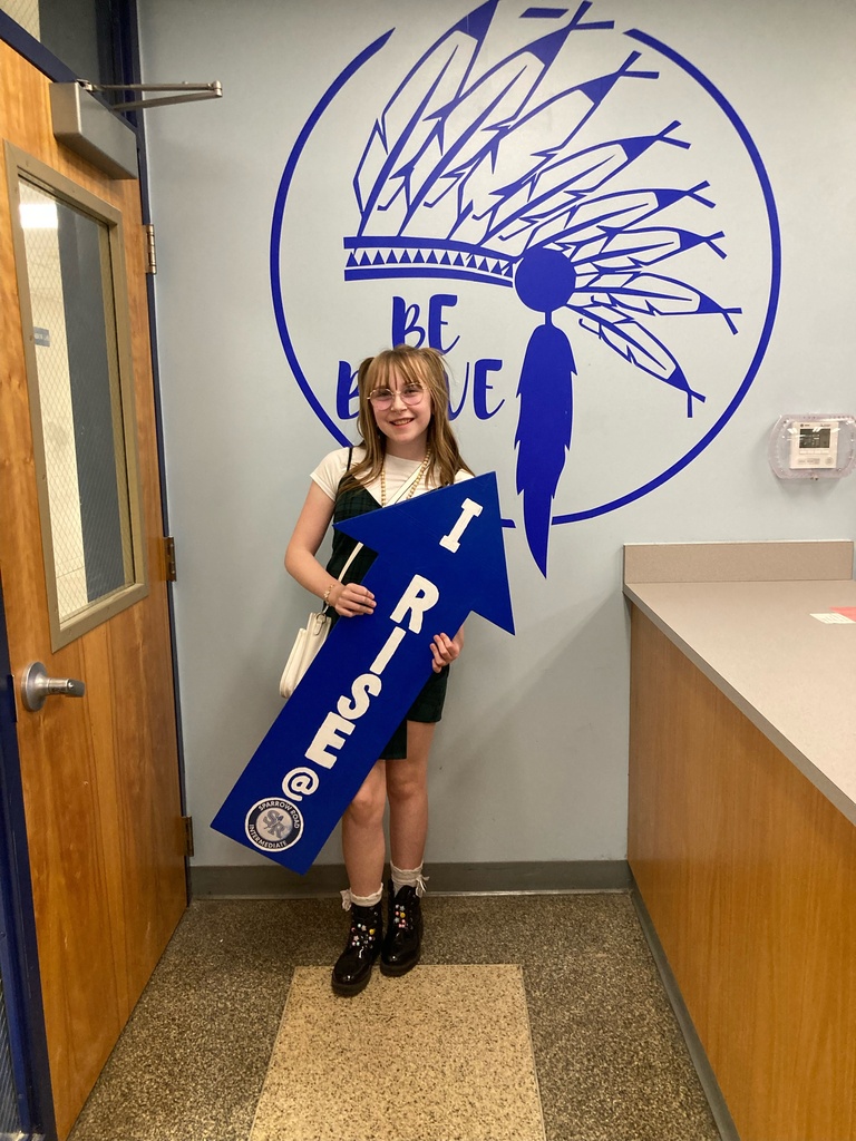 Student smiling and  standing in the main office proudly holding the I RISE sign.