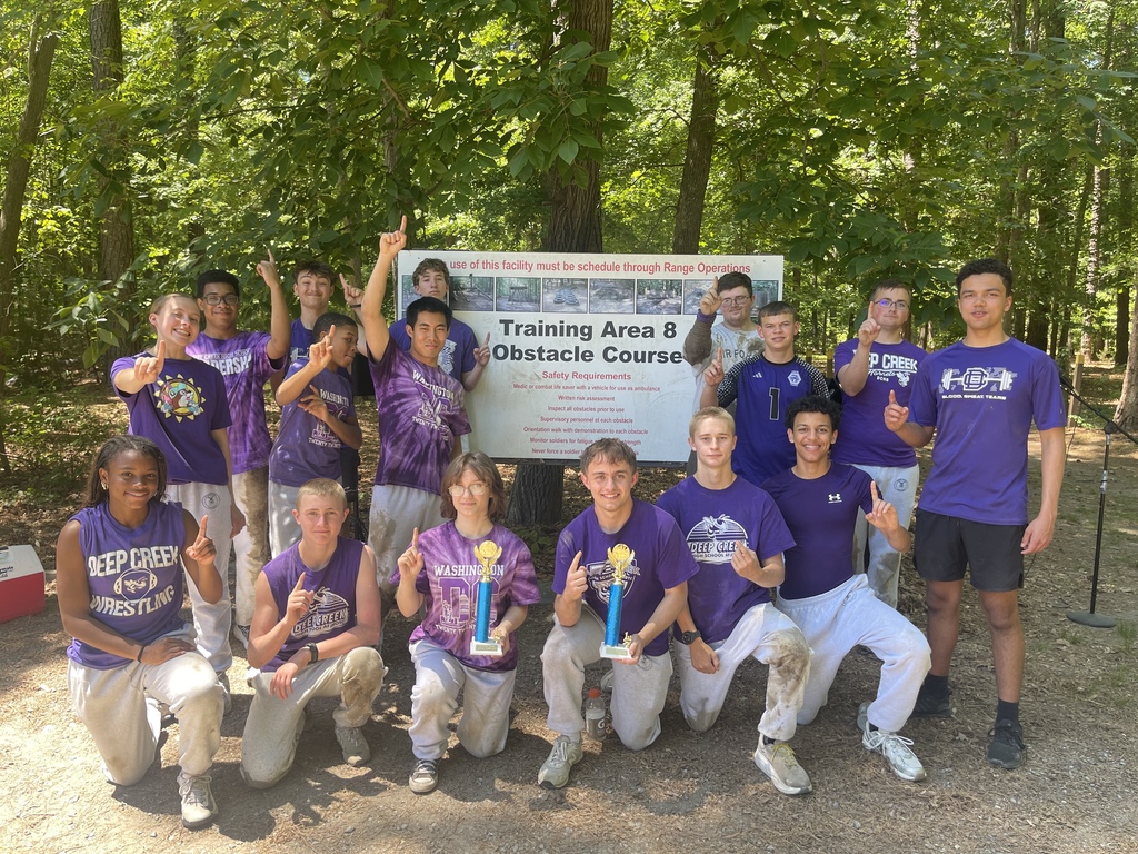 A group photo of fifteen high school students in purple athletic shirts celebrating outdoors in front of a "Training Area 8 Obstacle Course" sign. Two students in the front row hold trophies, and many point one finger up to signal "number one."
