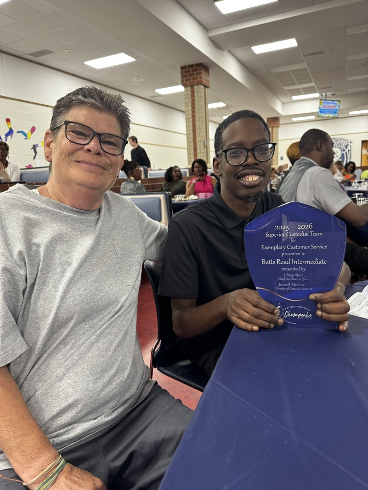 Two members of the Butts Road Intermediate custodial team sit together at the Chesapeake Public Schools custodial banquet while holding the school’s 2025–2026 Superior Custodial Team award for Exemplary Customer Service.