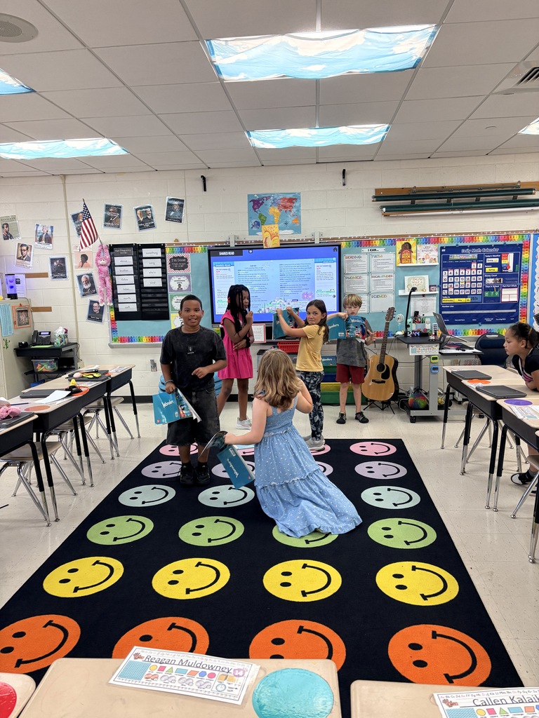 Classroom with six students performing a play five of the students, two boys and three girls are standing on a colorful smiley face carpet and one student is leaning on a student desk.