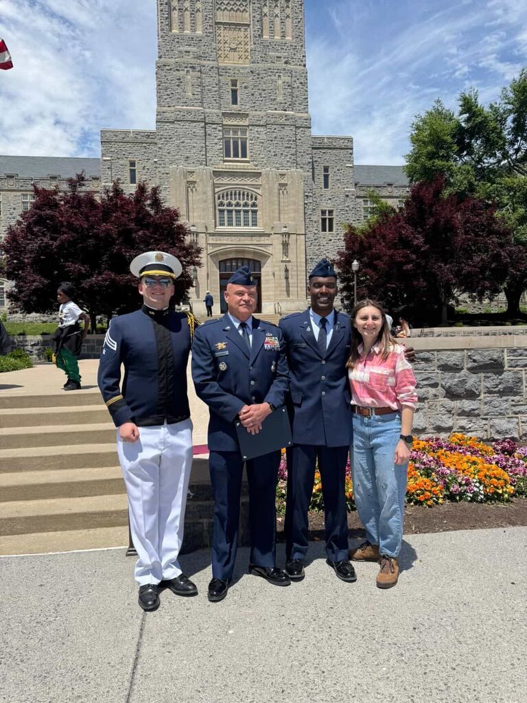 A group photo of four individuals standing on a concrete walkway in front of the stone facade of Burruss Hall at Virginia Tech. From left to right: a cadet wearing a dark navy dress jacket with white trousers, white service cap, and sunglasses; a senior military officer in a blue Air Force service dress uniform decorated with service ribbons and badges; a newly commissioned officer in a blue military uniform with a flight cap; and a young woman smiling, dressed in a pink and white plaid button-down shirt and light-wash blue jeans. Red trees and a colorful flower bed flank the group under a partly cloudy sky.