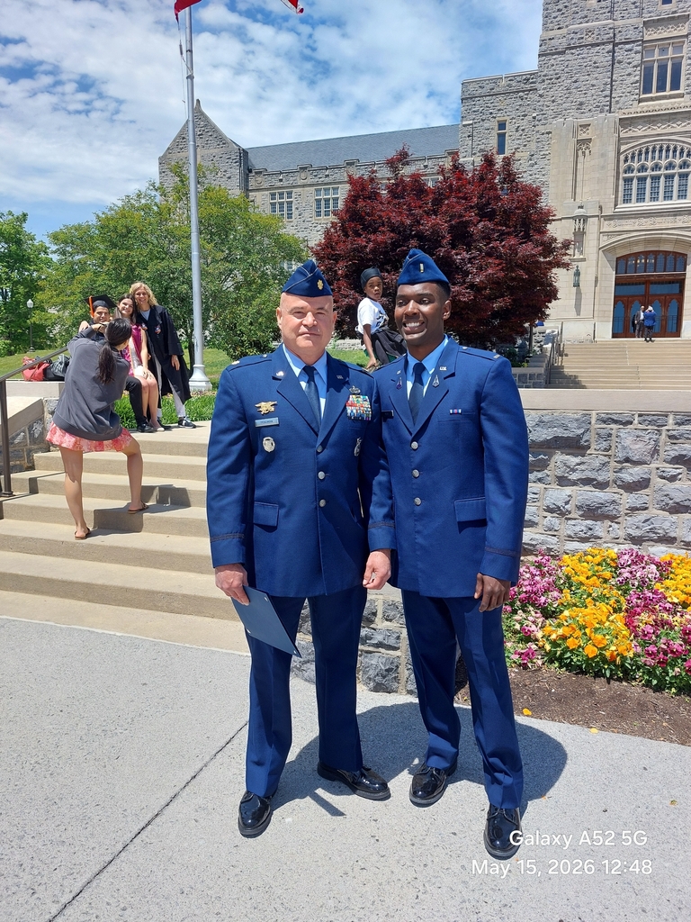 A full-length portrait of two military men standing together on a campus walkway with Virginia Tech's stone Burruss Hall in the background. On the left is an older officer in a blue service dress uniform adorned with numerous ribbons, badges, and insignia, holding a blue folder. On the right is a younger officer smiling, wearing a matching blue military dress uniform and flight cap with gold second lieutenant bars visible on his lapels. A vibrant flower bed with orange, purple, and yellow flowers is visible to the right, and other graduates are blurred in the background.