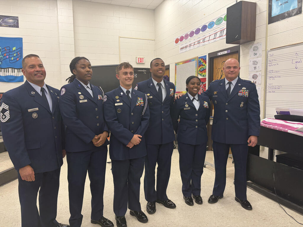 Six individuals in Air Force Junior ROTC (AFJROTC) blue service dress uniforms stand in a line side-by-side inside what appears to be a school music classroom. From left to right, there are four young men and two young women of diverse ethnicities, all smiling and posing for a group photo. The adult on the far right holds his arm around the shoulder of the young woman next to him. Their uniforms feature visible nametags, rank insignia, and colorful ribbon racks. The background includes a white brick wall, a whiteboard with musical notes, a door with an "EXIT" sign, and colorful music-themed posters.