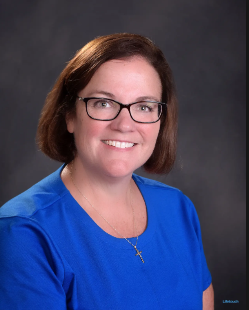 A professional studio headshot of a smiling woman with fair skin and shoulder-length brown hair. She is wearing black-rimmed glasses, a bright blue crewneck top, and a delicate gold chain necklace with a small cross pendant. She is posed against a dark, mottled gray background. A small "Lifetouch" watermark is visible in the bottom right corner.