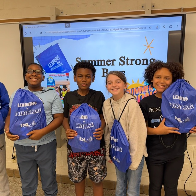 A group of elementary students smiling and holding up a special "Summer Strong" activity bags filled with books and learning games.