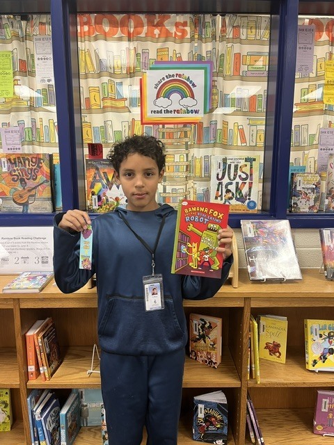 A smiling student proudly holds up their prize in the school library, celebrating their completion of the Read the Rainbow Book Challenge.