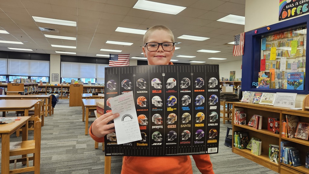 A smiling student proudly holds up their prize in the school library, celebrating their completion of the Read the Rainbow Book Challenge.