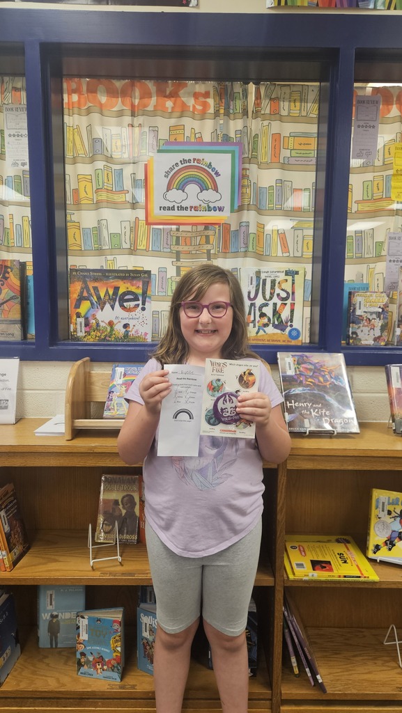 A smiling student proudly holds up their prize in the school library, celebrating their completion of the Read the Rainbow Book Challenge.