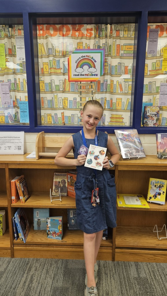 A smiling student proudly holds up their prize in the school library, celebrating their completion of the Read the Rainbow Book Challenge.