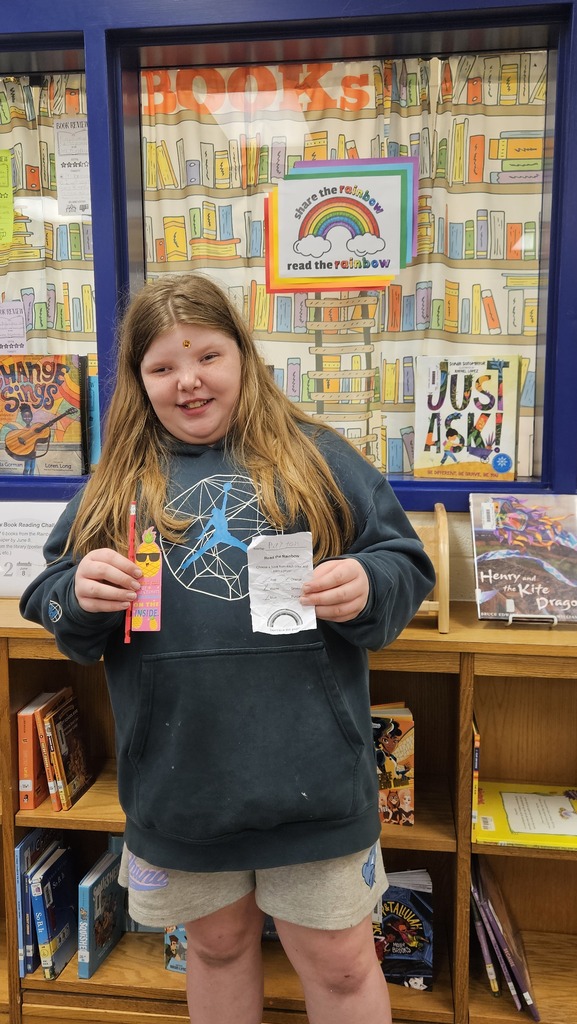 A smiling student proudly holds up their prize in the school library, celebrating their completion of the Read the Rainbow Book Challenge.