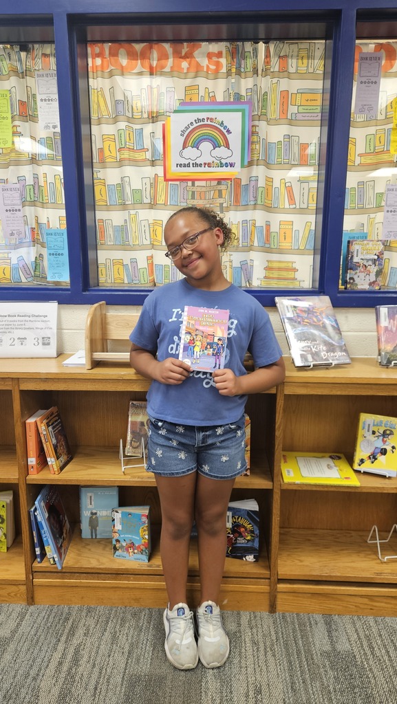 A smiling student proudly holds up their prize in the school library, celebrating their completion of the Read the Rainbow Book Challenge.