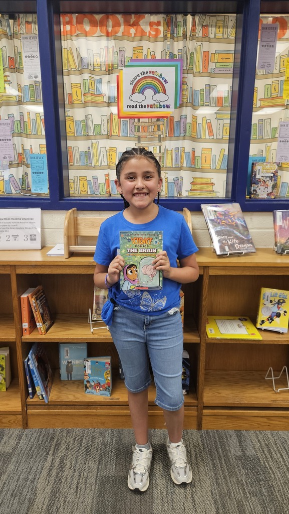 A smiling student proudly holds up their prize in the school library, celebrating their completion of the Read the Rainbow Book Challenge.