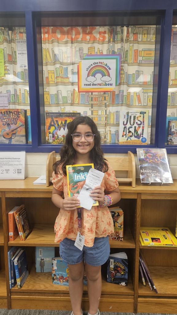A smiling student proudly holds up their prize in the school library, celebrating their completion of the Read the Rainbow Book Challenge.