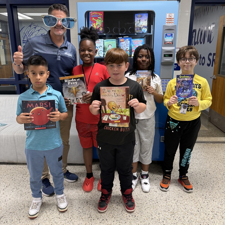A group of smiling students holing up their selected book with Mr. Triolet.