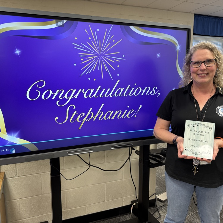 Mrs. Boyles smiling and holding her award plaque, standing next to the boxlight in the library. 