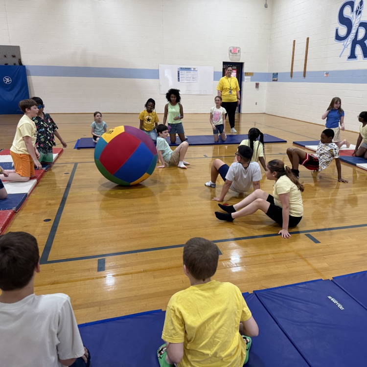 Students sitting in a square in the gym with a huge ball being pushed around. 