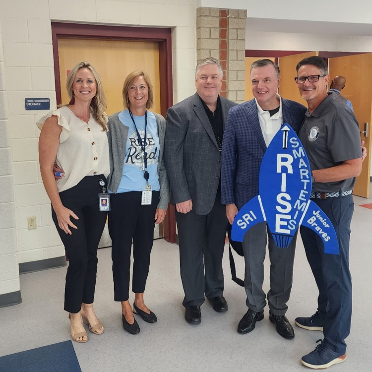 Wooden rocket cutout Smartemis posing with renowned psychologist Jodi Carrington, Superintendent Dr. Brubaker, Dr. Cotton, and School Board Chair Kim Scott with Mr. Triolet and Mrs. Boerboom at the Empowerment Conference.