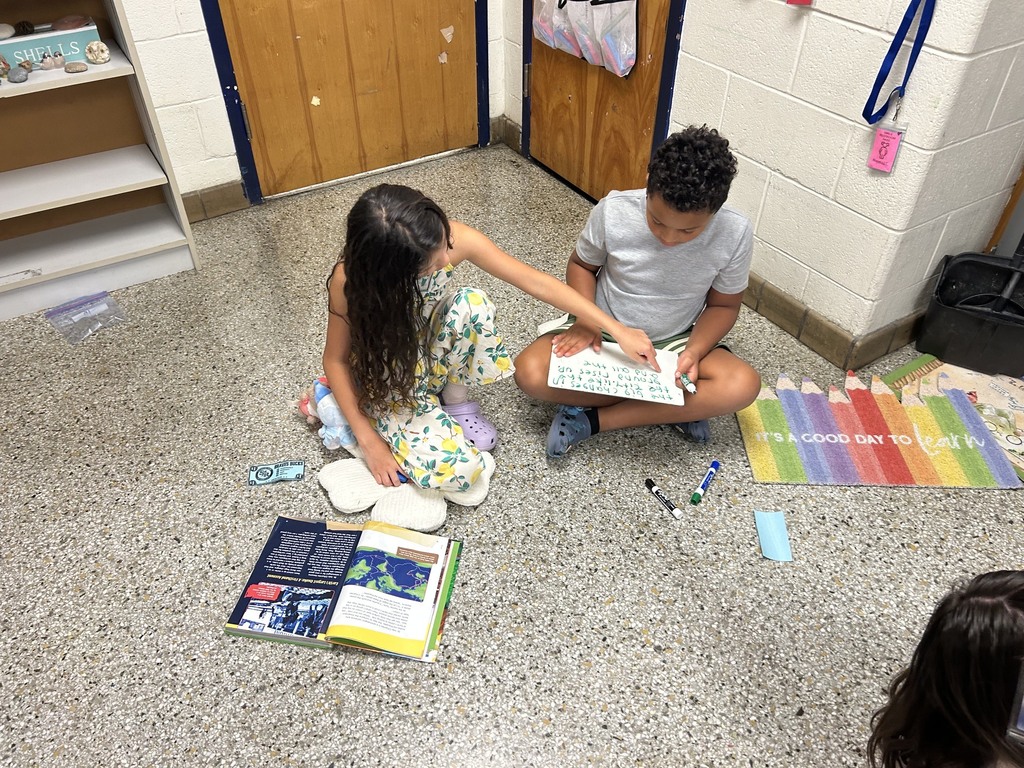 Two elementary students sit on the classroom floor reading a textbook together while writing their answers on a small whiteboard.