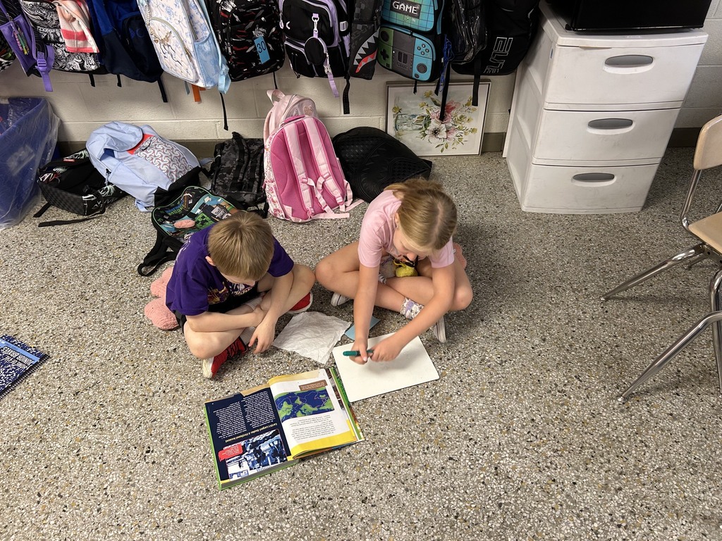 Two elementary students sit on the classroom floor reading a textbook together while writing their answers on a small whiteboard.