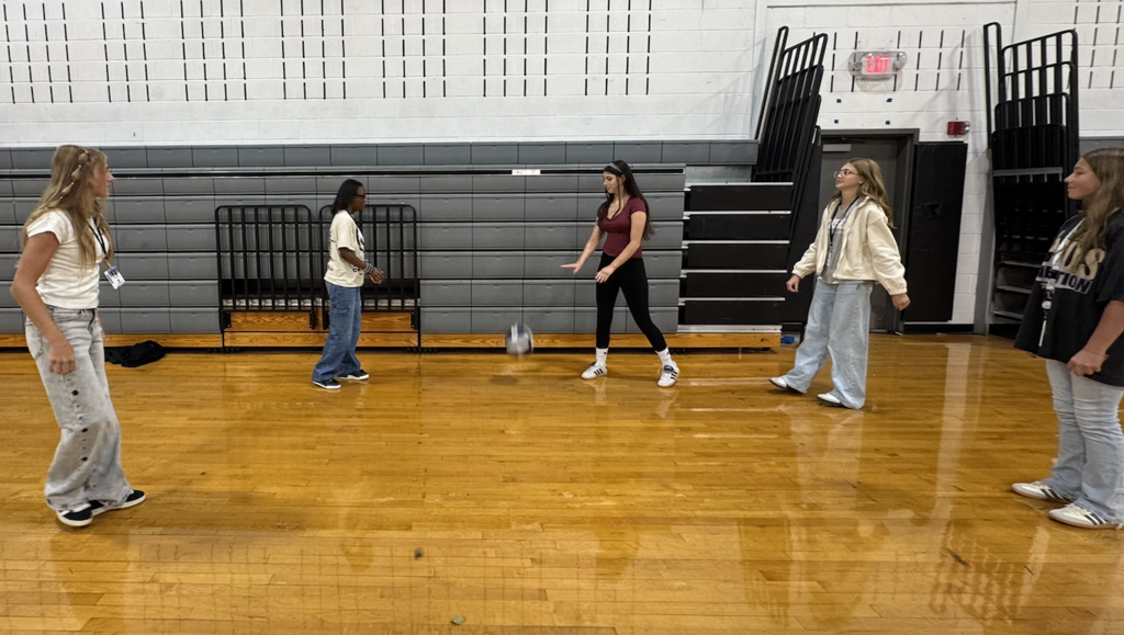 Five students standing in a circle playing volleyball indoors on a school gym wooden floor.