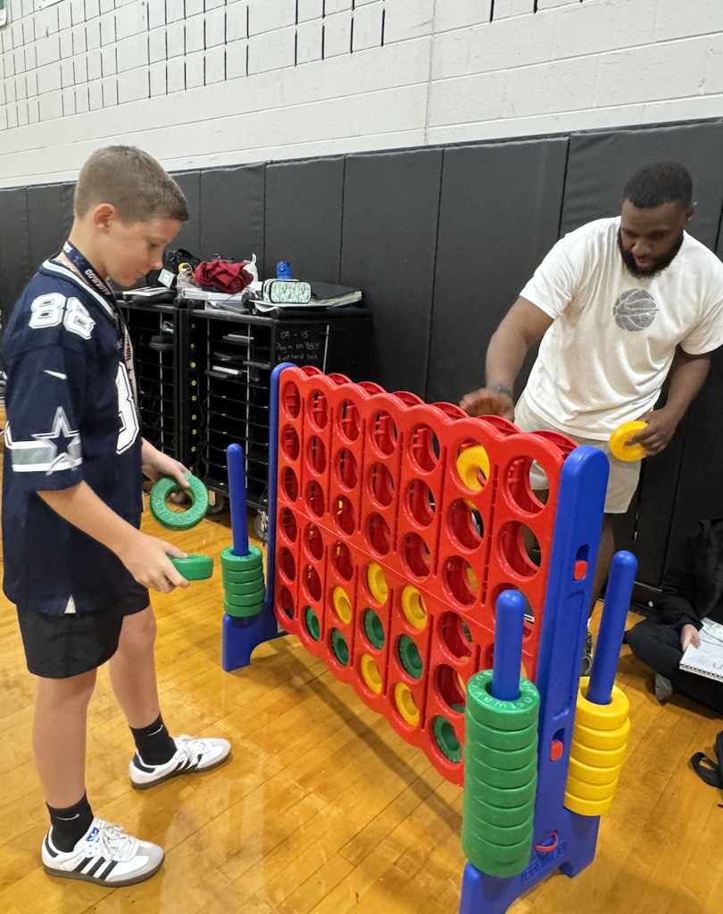 A young boy in a Dallas Cowboys jersey and an man play a large, life size Connect Four game indoors on a wooden gym floor.