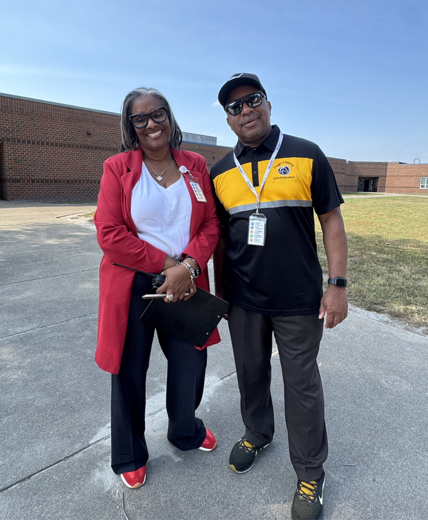 A woman in a red blazer and a man in a black and yellow polo shirt pose together outdoors in front of a school building.
