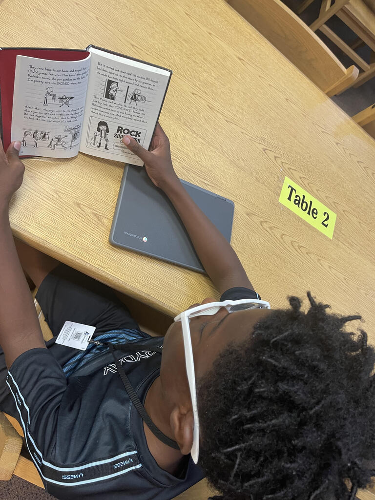 An overhead view of a student seated at a school desk reading an illustrated book.