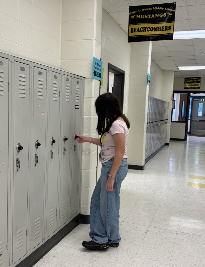 A student stands in a school hallway opening a grey metal locker.