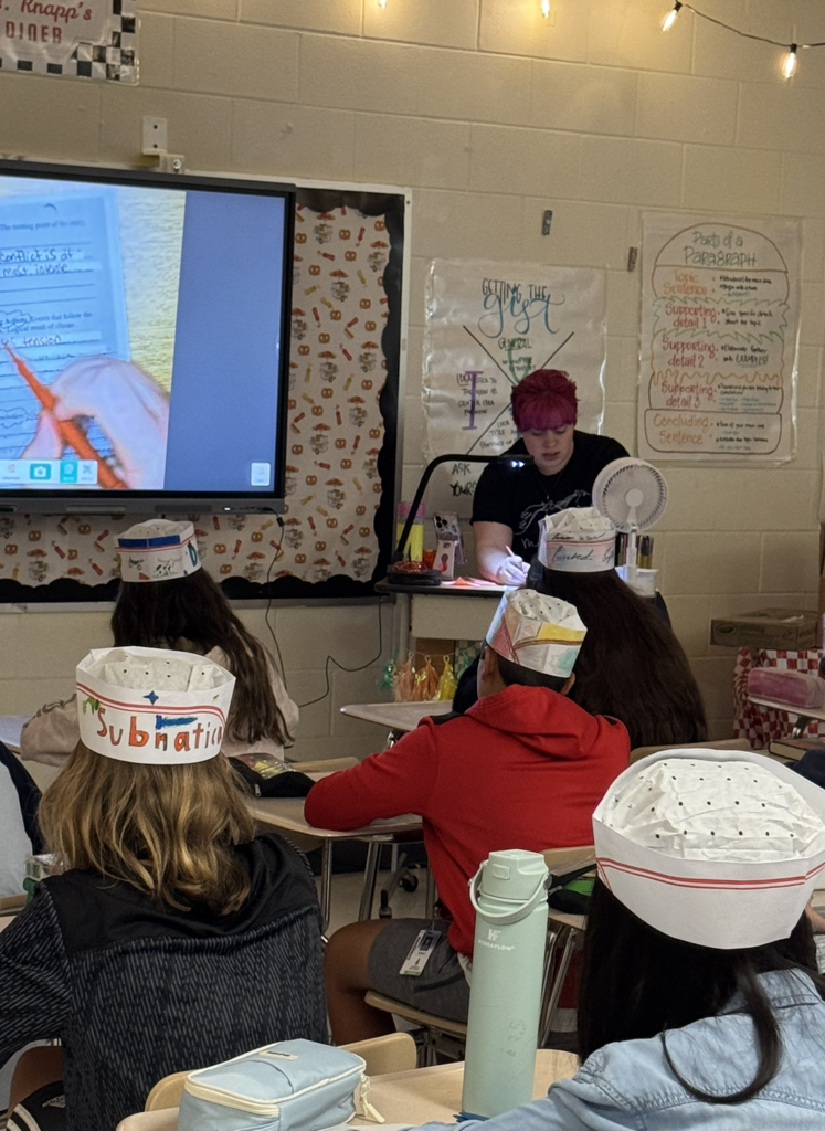 Students wearing paper diner chef hats sit at desks facing a teacher with short reddish hair in a classroom decorated with string lights and educational posters.