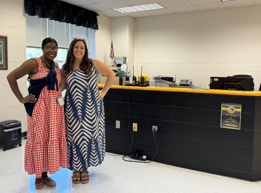 Two smiling women stand side by side in patterned maxi dresses in front of a school office reception desk.