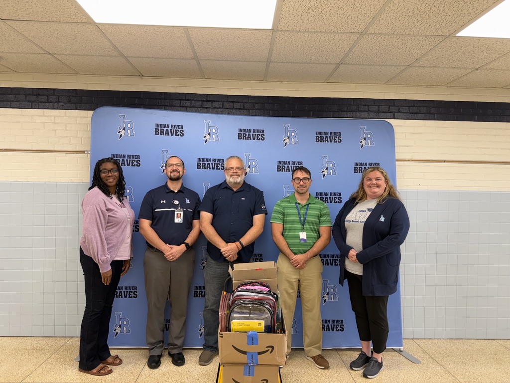 Five adults stand together in front of an Indian River Braves backdrop. In front of the group are boxes filled with donated school supplies, including backpacks and other classroom materials.