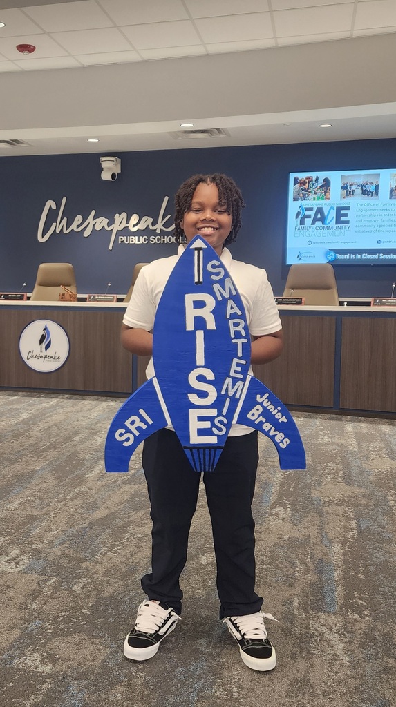 An SRI student  holding up a  "RISE / SMARTemis" rocket sign stands proudly in front of the Chesapeake Public Schools backdrop before a board meeting.
