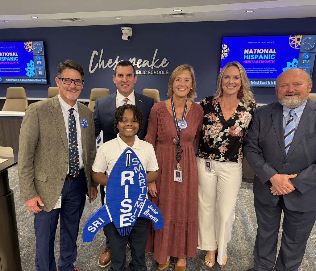 A student stands in the center of a group of school board members and SRI admin holding up a  "RISE / SMARTemis" rocket sign  in a school board meeting room.