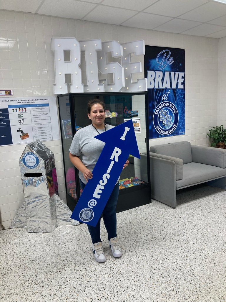 Teacher holding up the "I RISE" sign in the main lobby.