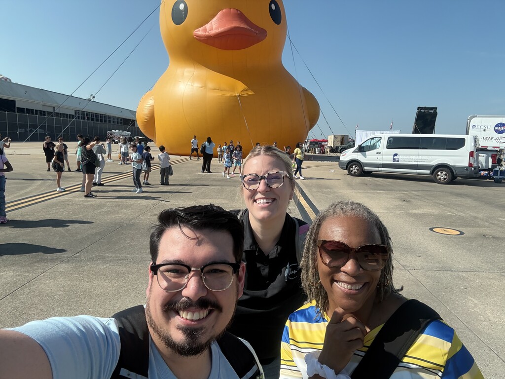 A selfie of three adults smiling outdoors on a tarmac, with a massive yellow rubber duck inflatable in the background.