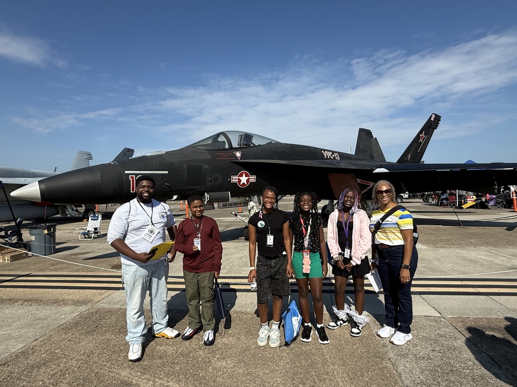 A group of six people—two adults and four kids—posing together outdoors in front of a large black military fighter jet on a sunny day.