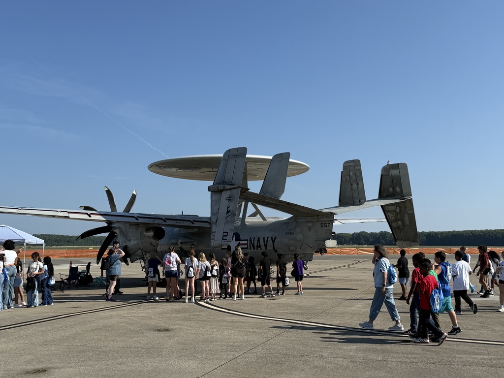 A large group of students and visitors gathered around a gray U.S. Navy E-2 Hawkeye radar aircraft parked on an airfield under a clear sky.