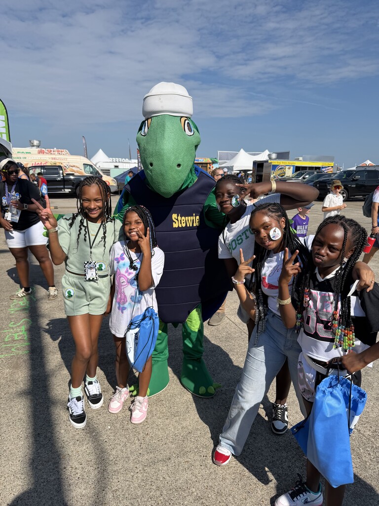A group photo of several young students posing cheerfuly outdoors alongside a green sea turtle mascot named "Stewie".