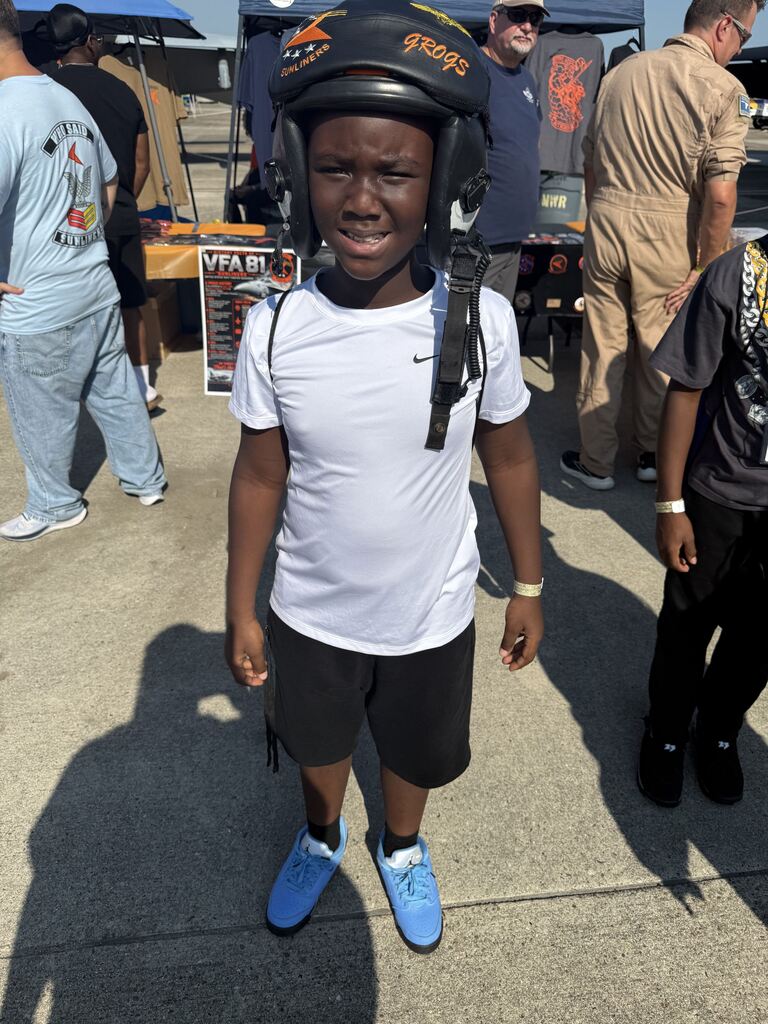 A young boy outdoors wearing a white t-shirt, black shorts, light blue sneakers, and a black military flight helmet labeled "FROGS".